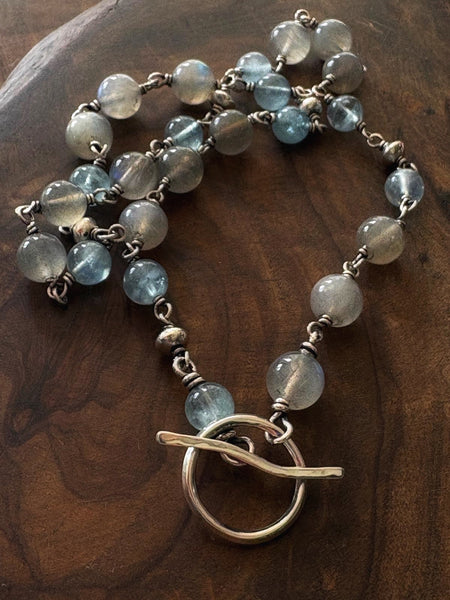 A beaded necklace featuring light blue labradorite and gray glass beads with an antique silver toggle clasp, displayed on a dark wood background.